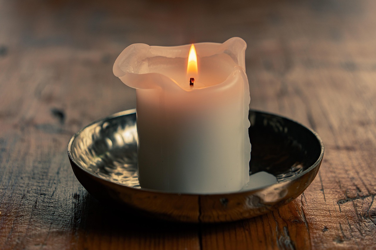 A burning white pillar candle with an uneven melted rim sits in a shallow metal dish on a wooden table.